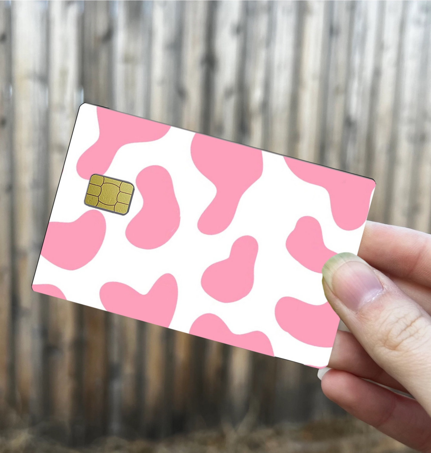 Cow print credit card skin in pink and white, with chip cutout, shown held in hand against wood background

