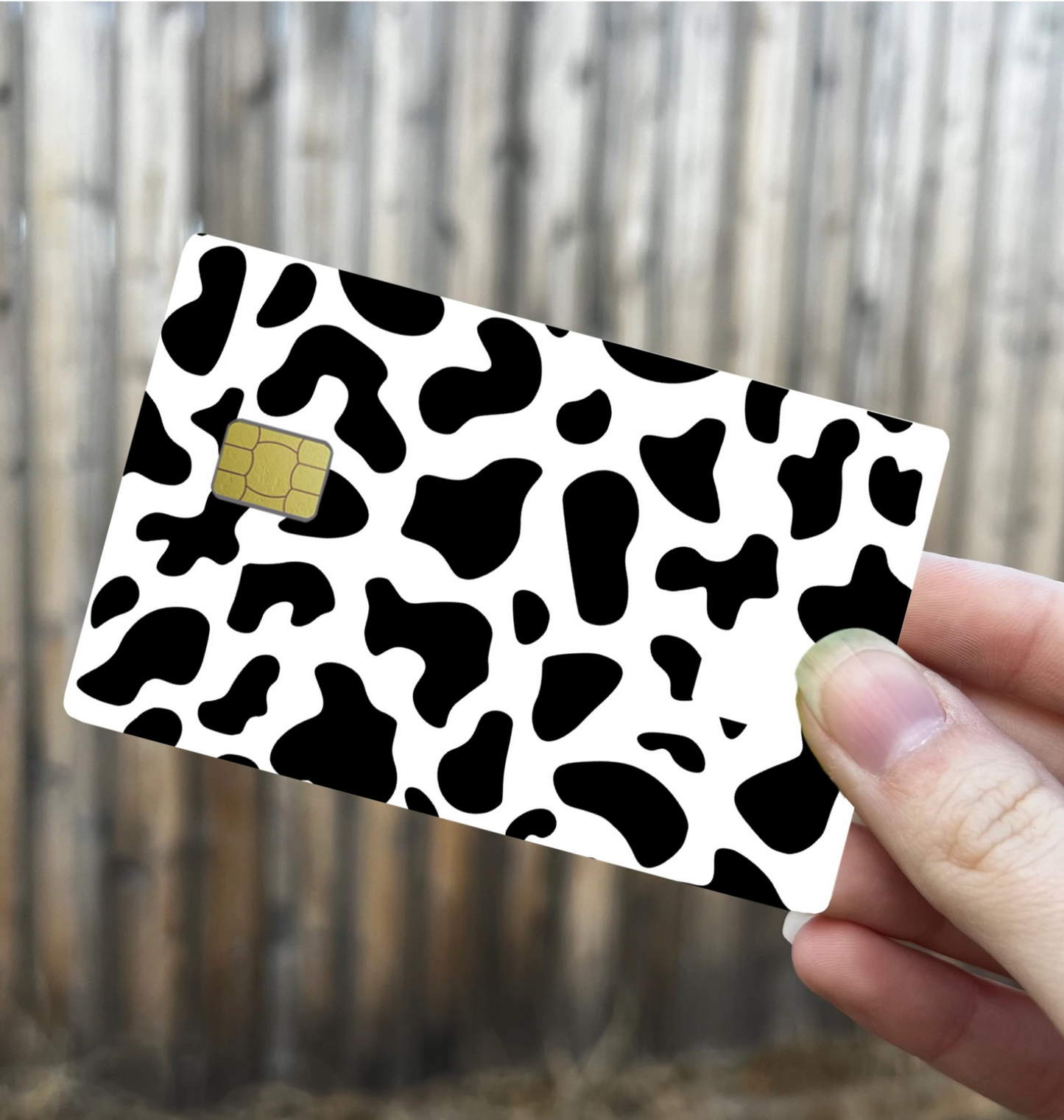 Cow print credit card skin in black and white, with chip cutout, shown held in hand against wood background
