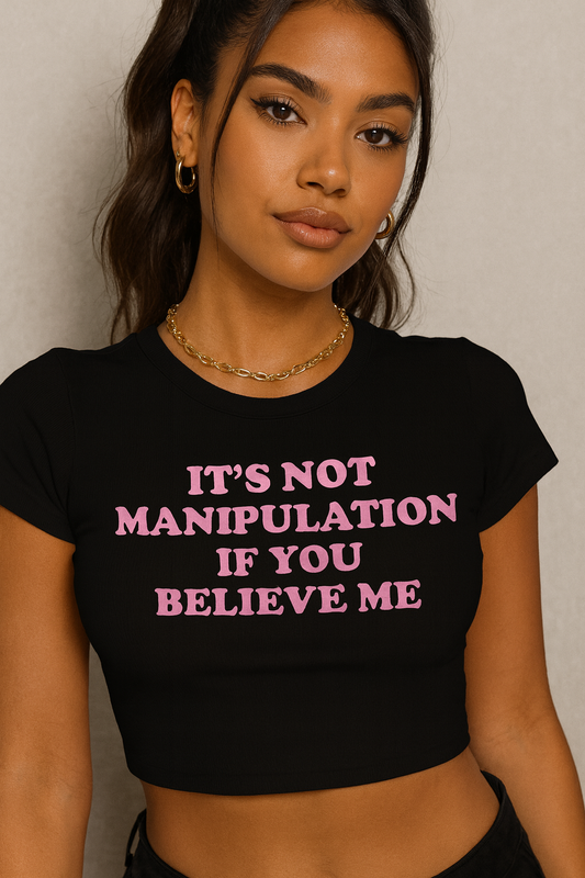 Young woman wearing a black crop top with pink bold text that says “It’s not manipulation if you believe me,” styled with gold hoops and chain necklace in confident, baddie aesthetic.