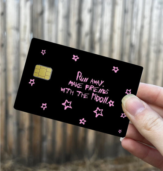 Black credit card skin with pink hand-drawn stars and text that says “Run away, make friends with the moon,” credit card size cover
