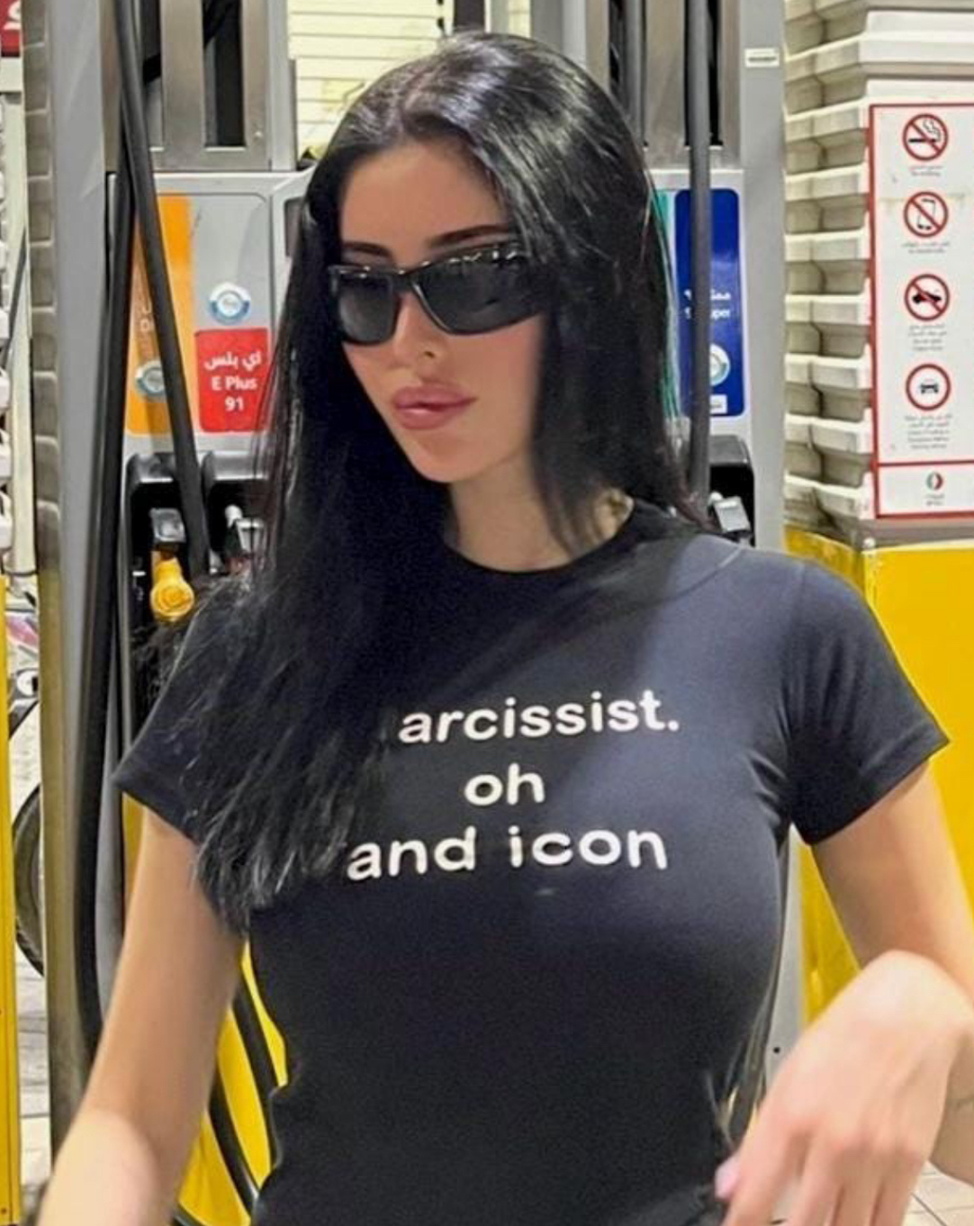 Woman in sunglasses wearing a fitted black graphic tee that says “narcissist. oh and icon.” standing at a gas station