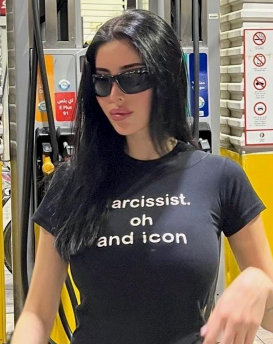Woman in sunglasses wearing a fitted black graphic tee that says “narcissist. oh and icon.” standing at a gas station